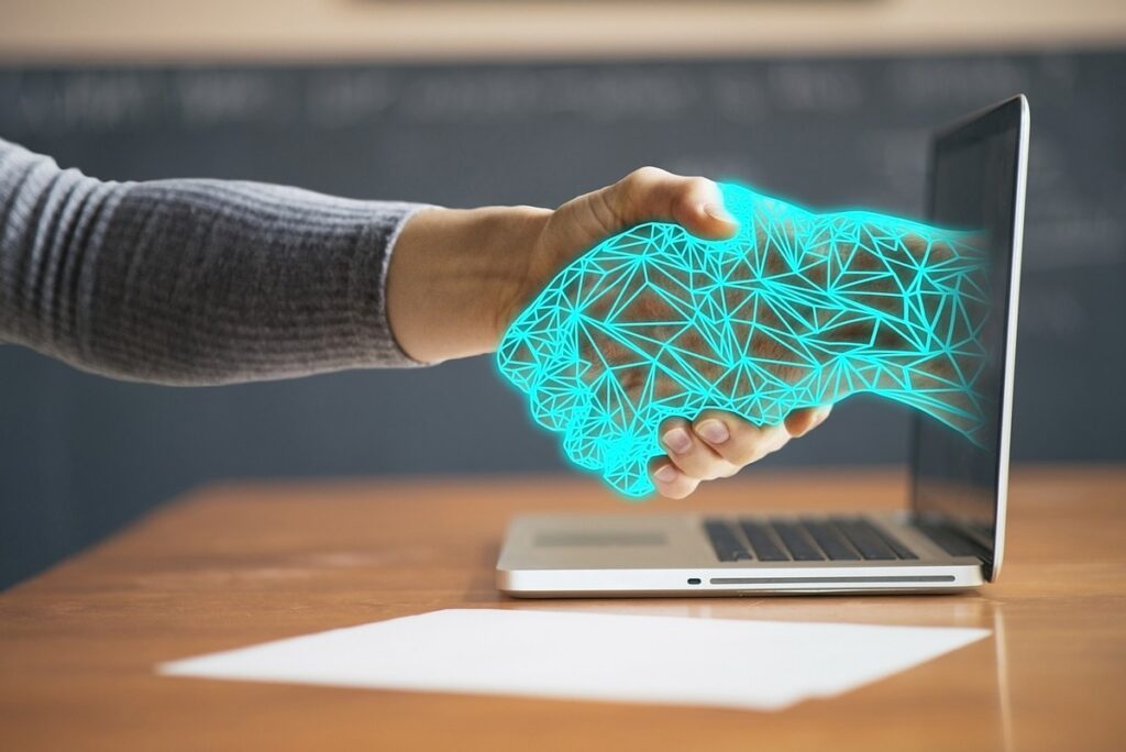 A human hand shakes with an AI technology hologram on a laptop screen